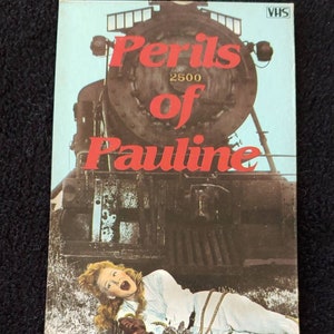 Puede incluir: Carátula de la cinta de video VHS para la película "Los peligros de Pauline" protagonizada por Betty Hutton, Billy DeWolfe, John Lund y William Demarest. La portada presenta una imagen en blanco y negro de una mujer atada frente a una gran locomotora de vapor.