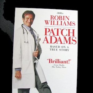 May include: A movie poster featuring Robin Williams as Patch Adams, a doctor who uses humour to help his patients. The poster has a white background with red text and a photo of Robin Williams in a white doctor's coat and red clown shoes.