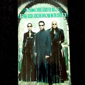 May include: A rectangular promotional card for the film The Matrix Reloaded featuring three figures in black attire and sunglasses standing before a stone archway with green digital code patterns cascading down the background and the title text above.