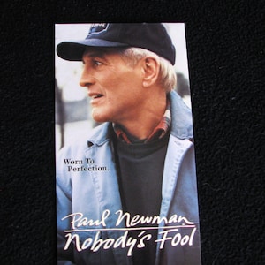 Könnte beinhalten: Eine Nahaufnahme von Paul Newman, der eine blaue Mütze und eine blaue Jacke trägt. Der Text "Worn To Perfection." befindet sich unter seinem Bild. Der Text "Paul Newman Nobody's Fool" befindet sich unten im Bild.
