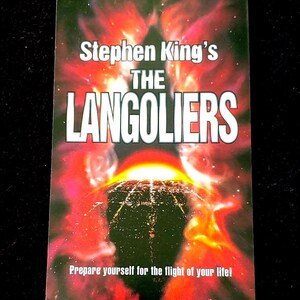 Op de afbeelding: Een rechthoekige boekomslag met een levendige rode en oranje nevelachtergrond. De tekst luidt Stephen King's THE LANGOLIERS in witte en rode letters. Daaronder bevindt zich een donker landschap met een rasterpatroon onder een fel licht. Onderaan staat de tekst Prepare yourself for the flight of your life!