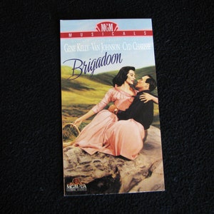 May include: A VHS tape cover for the musical film Brigadoon, featuring Gene Kelly, Van Johnson, and Cyd Charisse. The cover shows a romantic scene with the couple embracing in a green field.