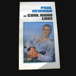 Könnte beinhalten: Ein Vintage-Filmplakat mit Paul Newman als Cool Hand Luke. Das Plakat ist weiß mit schwarzem Text und einem Foto von Paul Newman in einem blauen Hemd, der sitzt und eine Zigarette raucht.