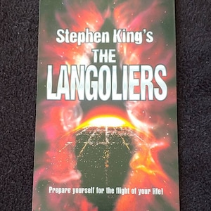 May include: A book cover for Stephen King's novel "The Langoliers". The cover features a fiery red and orange background with a dark, circular object in the center. The text "Stephen King's The Langoliers" is in white letters at the top of the cover. The text "Prepare yourself for the flight of your life!" is in white letters at the bottom of the cover.