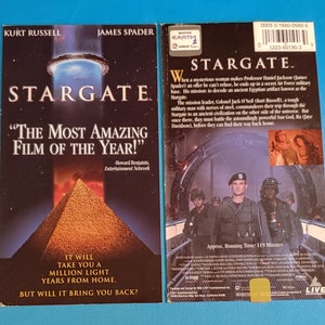 Puede incluir: Una carátula de VHS para la película Stargate, con Kurt Russell y James Spader. La carátula tiene un fondo negro con una gran imagen de la puerta de las estrellas, un portal circular, en el centro. El texto "Stargate" está en letras blancas grandes en la parte superior de la carátula. El texto "¡La película más increíble del año!" está en letras blancas debajo de la puerta de las estrellas. La carátula también presenta una cita de Howard Benjamin, Entertainment Network.