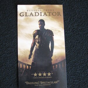 May include: A movie poster for the film Gladiator starring Russell Crowe. The poster features a close-up of Crowe as Maximus Decimus Meridius, a Roman general, in full armor. The poster has a dark background with a bright sky in the background. The text on the poster reads "RUSSELL CROWE GLADIATOR".