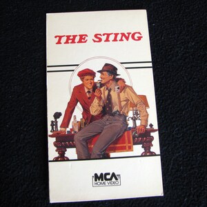 Könnte beinhalten: Ein Vintage-VHS-Kassettencover für den Film "The Sting" mit zwei Männern im Anzug, einer mit rotem Hut, die an einem Tisch mit einem Getränk sitzen. Das Cover trägt das Logo von MCA Home Video.