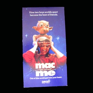 May include: A promotional poster for the film "Mac and Me" features a whimsical alien character atop a person's head. The poster has the text "How two boys worlds apart become the best of friends." and "Out of this world and into your heart."