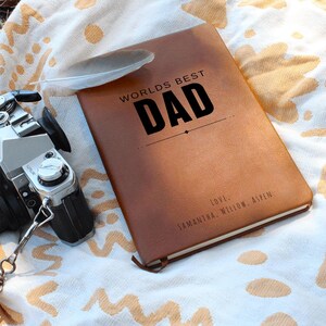 Puede incluir: Un diario de cuero marrón con el texto "WORLD'S BEST DAD" en negro. El diario tiene una cinta de marcador y una pluma encima. Las palabras "LOVE. SAMANTHA, WILLOW, ASPEN" están impresas en la parte inferior.