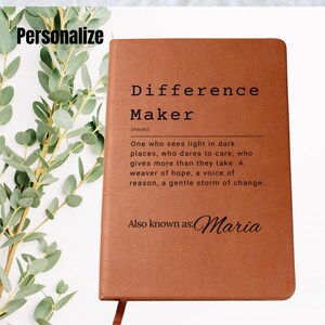 Puede incluir: Un diario de cuero marrón con el texto "Difference Maker" y una definición de la palabra. El texto "También conocido como: Maria" está impreso debajo de la definición.