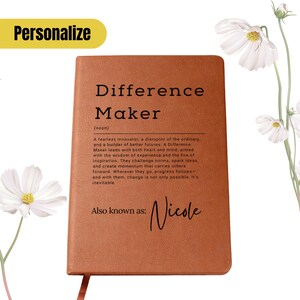Puede incluir: Un cuaderno de cuero marrón con las palabras "Difference Maker" y una definición impresa en la portada. El nombre "Nicole" también está impreso en la portada. El cuaderno tiene una cinta de marcador.