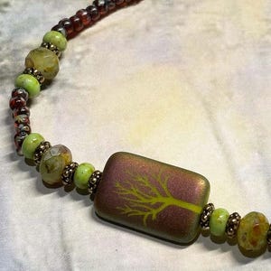 May include: A beaded necklace featuring a rectangular pendant with a lime green tree design on a burgundy background. The necklace includes green, brown, and gold-colored beads. The beads are strung together to create a unique and artistic piece of jewelry.