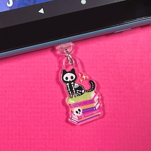 May include: A clear acrylic keychain featuring a skeleton cat sitting atop a stack of colorful books. The cat has a black body with white bones and a star above its head. The books are pink, purple, and green. The keychain is attached to a device.