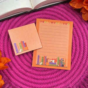 May include: Orange notepad and sticky note set with a fall theme. The notepad has lined pages, and both feature illustrations of books, pumpkins, and a fox. The set is on a pink woven placemat with a book and orange flowers in the background.