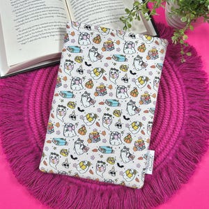 May include: A white book sleeve with a repeating pattern of cartoon ghosts, books, pumpkins, and bats. The sleeve is resting on a pink fringed placemat next to an open book and a small potted plant. The background is a solid bright pink.