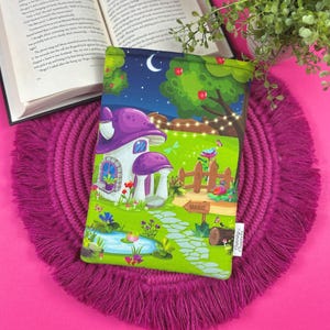 May include: A colorful book sleeve featuring a whimsical mushroom house scene with a night sky, trees, and a path leading to a sign that says "MAGIC". The sleeve is on a pink fringed placemat with an open book in the background.