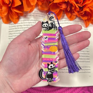 Puede incluir: Un marcapáginas con temática de Halloween con una pila de libros, una calabaza y un diseño de calavera. El marcapáginas presenta un diseño de esqueleto de gato negro y una borla morada. El fondo incluye flores naranjas y un libro.