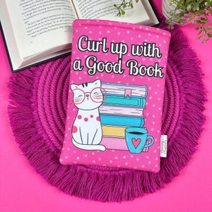 Peut inclure: Une housse de livre rose avec le texte "Curl up with a Good Book" et un chat de dessin anim&eacute; portant des lunettes. Le motif comprend une pile de livres, une tasse &agrave; caf&eacute; et de petits c&oelig;urs. La housse est sur un dessous de plat rose &agrave; franges.