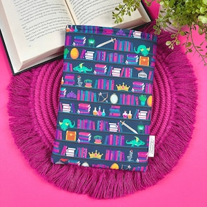 Puede incluir: Una funda para libros de color azul oscuro con un patrón repetido de libros, coronas, pociones y dragones. El texto "Charming" está impreso en la funda.