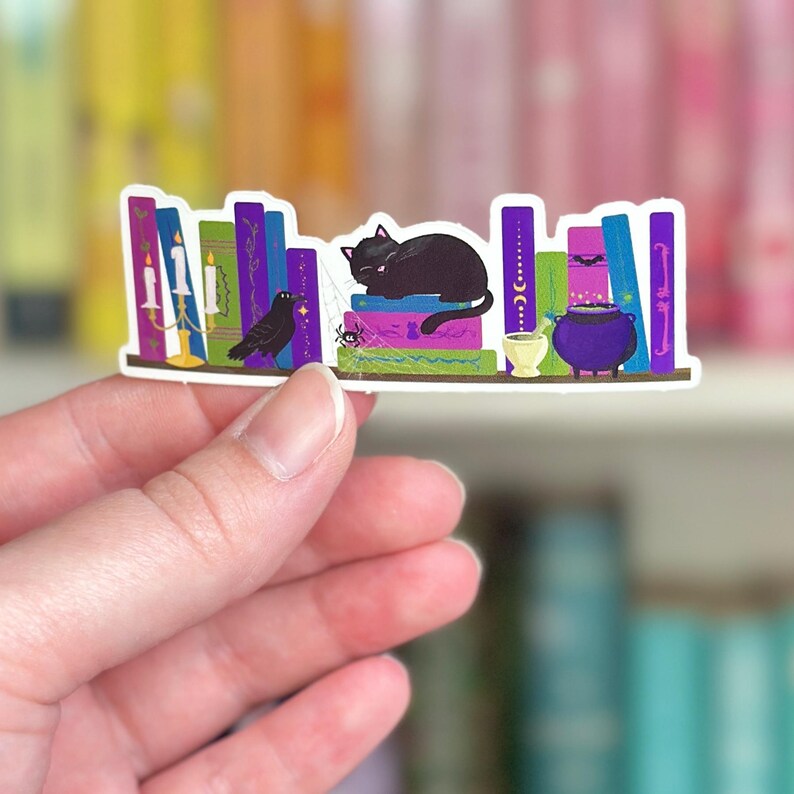 May include: A sticker of a bookshelf with a black cat sleeping on top of a stack of books. There is a raven, a spider, candles, a cauldron, and a mortar and pestle on the shelf. The sticker is on a white background.