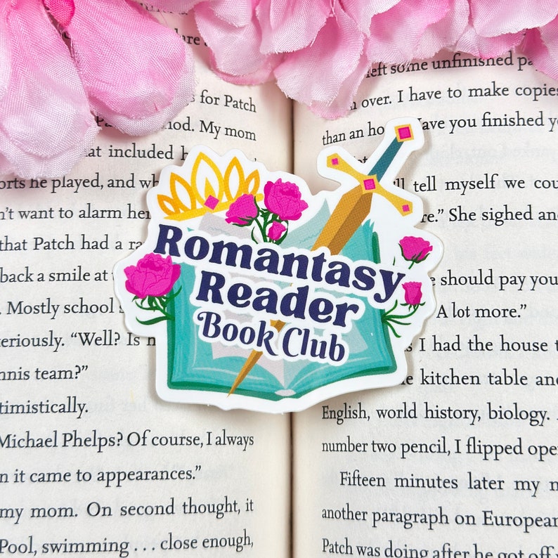May include: A sticker with a blue book, a sword, and pink flowers. The text on the sticker reads "Romantasy Reader Book Club".
