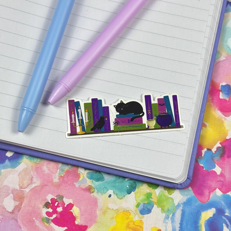 May include: A sticker featuring a black cat sitting on a bookshelf with a variety of books, a black bird, a candle, and a cauldron. The books have purple, green, and gold spines with text on them.