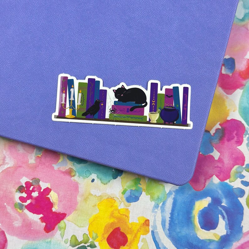 May include: A black cat sleeps on a shelf of books with a black bird, candles, and a cauldron. The books are purple, green, and blue with gold accents. The shelf is white.