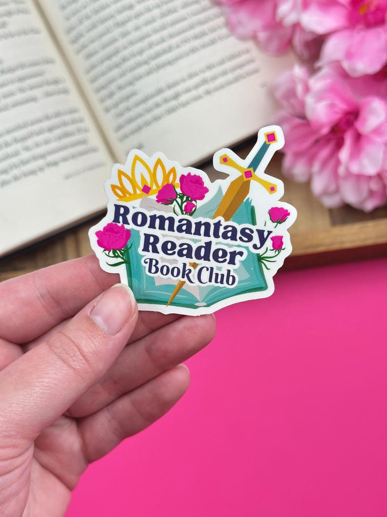 Hand holding a vinyl sticker that has a fantasy design that says "romantasy reader book club", with an open book and flowers in the background.
