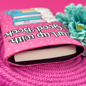 Peut inclure: Une housse de livre rose avec un motif de c&oelig;urs blancs et le texte "Curl up with a good book". La housse pr&eacute;sente une illustration color&eacute;e de livres et d'une licorne. Un livre est partiellement ins&eacute;r&eacute;.