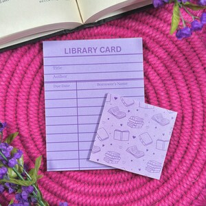 May include: A light purple library card and a small square notepad with a book pattern. The library card has fields for title, author, due date, and borrower's name. The background is a bright pink woven surface.