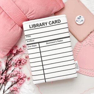 May include: A white and black library card-themed book sleeve with lines for title, author, due date, and borrower's name. The text "LIBRARY CARD" is at the top. The background includes pink items and floral accents.