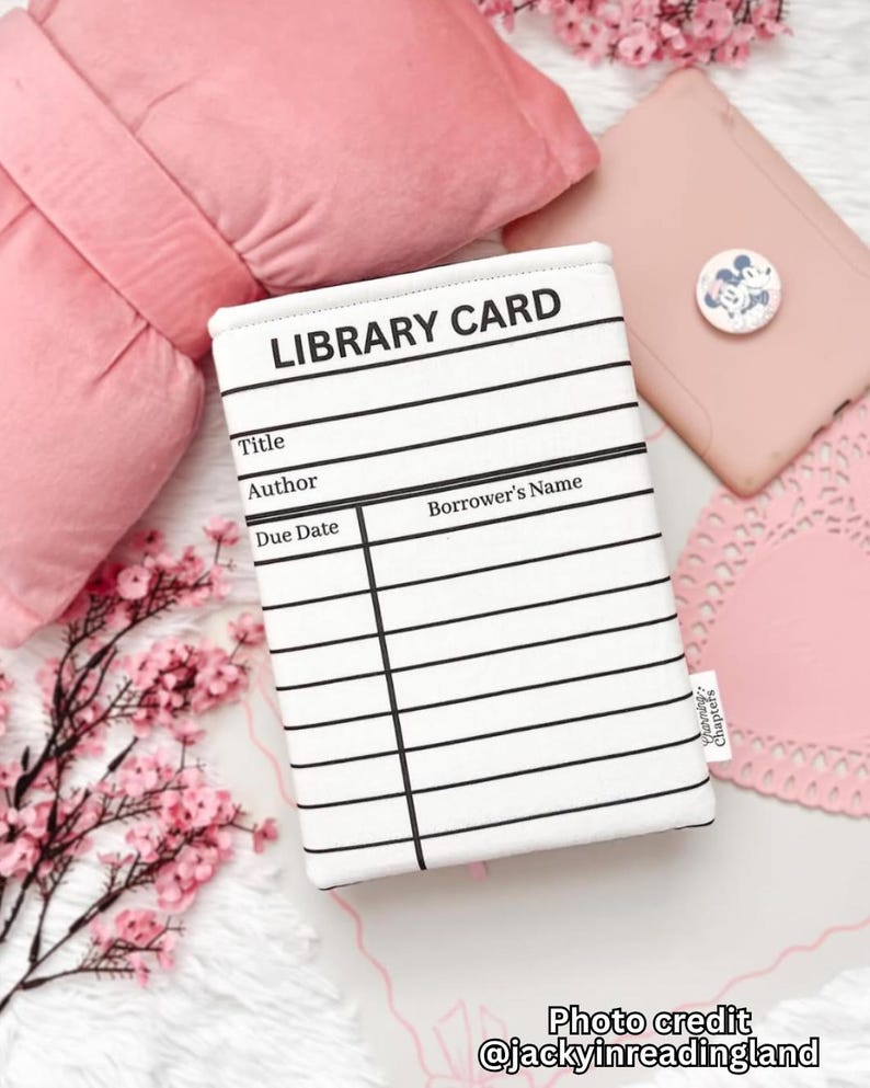 May include: A white and black library card-themed book sleeve with lines for title, author, due date, and borrower's name. The text "LIBRARY CARD" is at the top. The background includes pink items and floral accents.