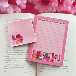 May include: Two pink notepads with a heart border and a shelf design with books, a teddy bear, and plants. The notepads are on an open book, with pink flowers in the background. Measurements are not applicable.