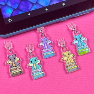 May include: Five colorful dragon book charms with a stack of books. The dragons are purple, blue, green, yellow, and gray. The charms are made of clear acrylic and are attached to a clear plastic string.