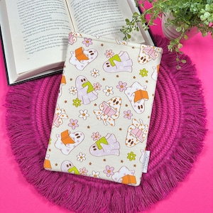 May include: A cream-colored book sleeve featuring a repeating pattern of ghosts, books, and flowers in shades of purple, orange, and green. The sleeve rests on a pink fringed placemat next to an open book and a small potted plant.