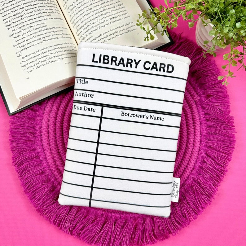 Foam padded book cover laying flat next to an open book.  The book sleeve has a design made to look like a library card.   It is great to take to book signings and events to collect author signatures.