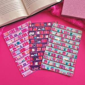 May include: Three decorative book-themed notebooks with colorful bookshelf designs. The notebooks feature pink, purple, and blue backgrounds with illustrations of books, cats, and decorative items. The word "LOVE" is printed on two of the notebooks.