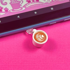 May include: A white ceramic mug with a brown coffee drink and latte art. The mug is attached to a clear plastic charm.