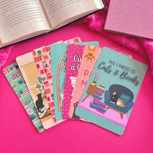 May include: A collection of colorful cat-themed bookmarks with various designs, including cats, books, and the text "All I Need Is Cats & Books." The bookmarks are arranged on a vibrant pink surface, alongside an open book and a pink glitter notebook.