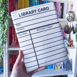 May include: A white book sleeve with a black library card design. The text "LIBRARY CARD" is at the top, with lines for title, author, and borrower's name. The background shows a bookshelf filled with colorful books.