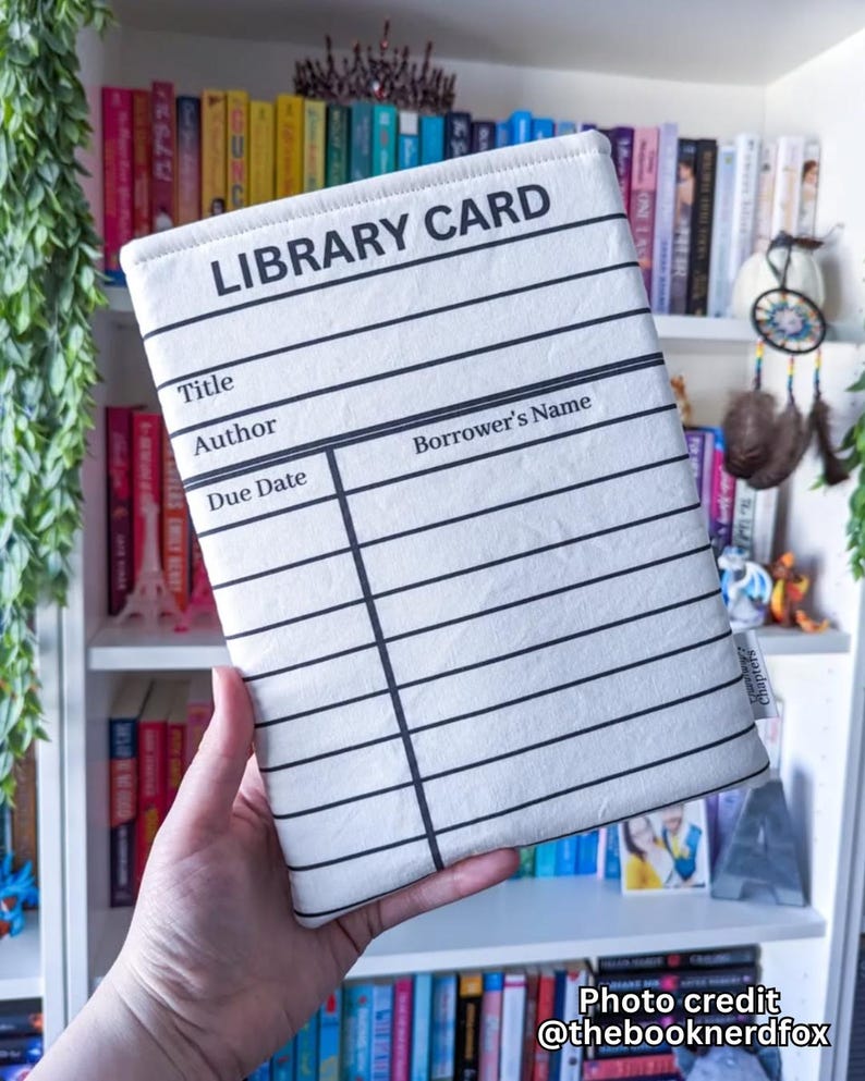 May include: A white book sleeve with a black library card design. The text "LIBRARY CARD" is at the top, with lines for title, author, and borrower's name. The background shows a bookshelf filled with colorful books.