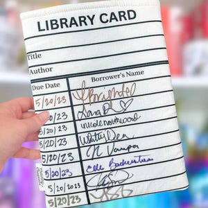 May include: A white library card sleeve with black lines and text. The text "LIBRARY CARD" is at the top. The sleeve has sections for title, author, due date, and borrower's name. Handwritten names and dates fill the spaces.