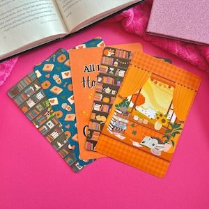 May include: A collection of colorful, illustrated bookmarks with cozy, book-themed designs. One bookmark reads "All I Have is Hot Tea". The bookmarks are arranged on a bright pink surface, near an open book and a pink glitter notebook.