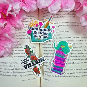 May include: Four colorful stickers featuring book-themed illustrations. The first sticker is a teal and white sticker with a dragon sitting on a stack of books. The second sticker is a teal and white sticker with the text "Romantasy Reader Book Club". The third sticker is a red, white, and black sticker with a sword and roses. The fourth sticker is a red, white, and black sticker with the text "Team whoever the Villain is..."
