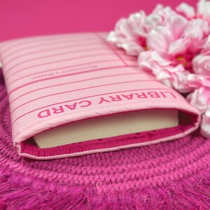 May include: A pink book cover with the words "LIBRARY CARD" printed on it. The cover is made of a soft, fabric-like material and has a white book spine.