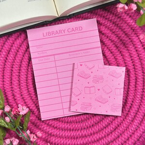 May include: A pink library card and a small notepad with a book pattern. The library card has lines for title, author, due date, and borrower's name. The notepad features a repeating pattern of books and hearts.