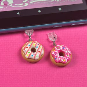 May include: Two donut-shaped phone charms, one with pink and white frosting and sprinkles, the other with pink frosting and a white swirl design.