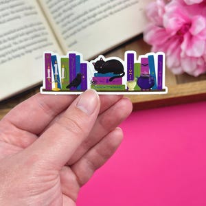 Hand holding a sticker with a dark academia bookshelf design that includes a black cat, cauldron, raven and candelabra. The sticker is being held over a pink background with an open book and pink flowers.