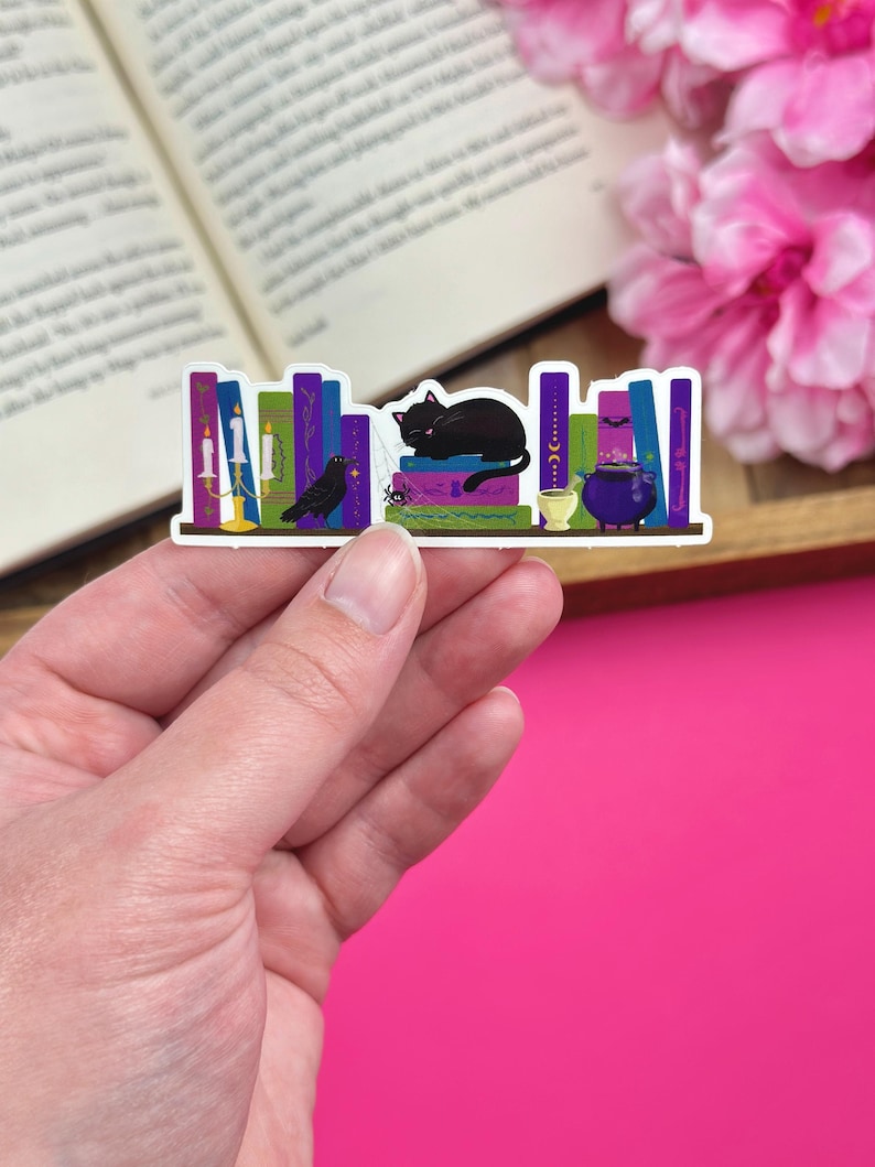 Hand holding a sticker with a dark academia bookshelf design that includes a black cat, cauldron, raven and candelabra. The sticker is being held over a pink background with an open book and pink flowers.