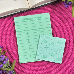May include: A mint green library card notepad and a small square sticky note with a book pattern. The library card notepad has lines for title, author, due date, and borrower's name. The items are on a pink woven mat.